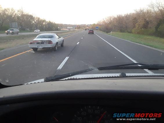 White 1980s Chevrolet Camaro driving on highway, rear view