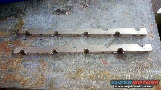 Two identical, long, flat, metallic engine parts with circular cutouts and small holes, lying on a textured metal surface