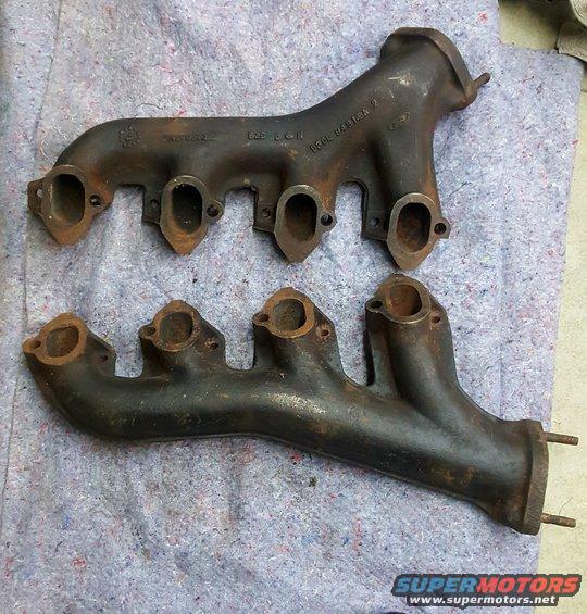Two cast iron exhaust manifolds, one above the other, on a grey cloth surface. Text is visible on the upper manifold