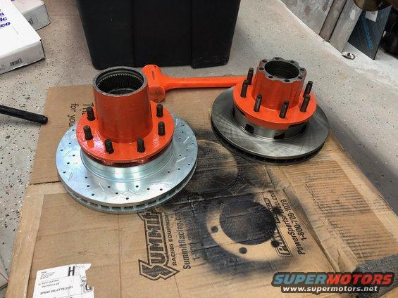 Two orange-painted wheel hubs with brake rotors underneath, sitting on a cardboard box. A sledgehammer is nearby