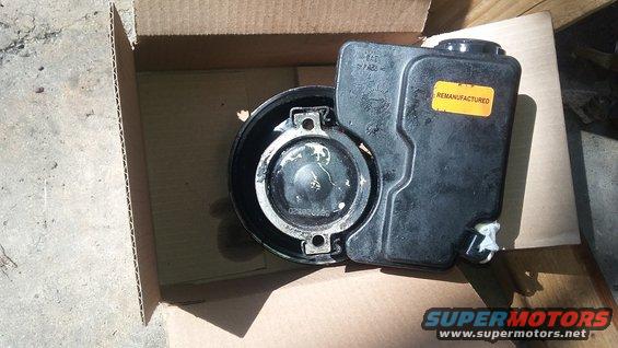 Black plastic power steering fluid reservoir with a yellow "REMANUFACTURED" sticker, inside a cardboard box