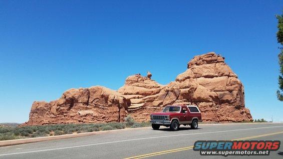 Red Ford Bronco driving on a road with large red rock formations in the background