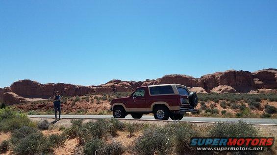 1985 Ford Bronco parked on the side of a desert highway with red rock formations in the background