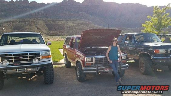 Red Ford Bronco with hood open, parked next to a white Ford truck and a dark blue Ford Bronco