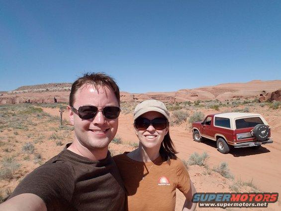 Two people smile for a selfie with a red 1980s Ford Bronco parked in a desert landscape