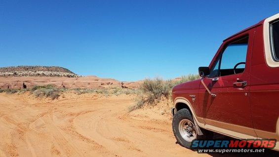 Red and tan Ford Bronco parked on a dirt road in a desert with red rock formations under a clear blue sky