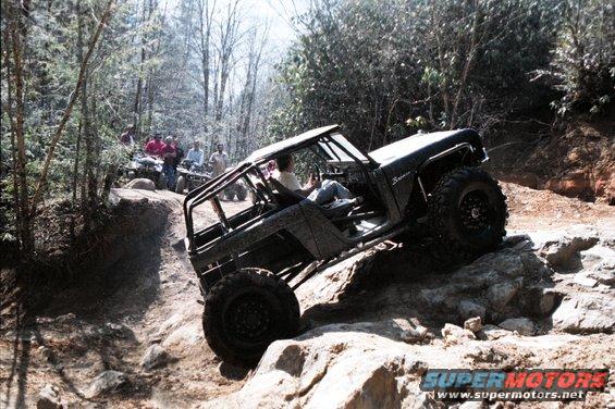 Black 1976 Ford Bronco climbing rocks on a wooded trail, with people on ATVs in the background