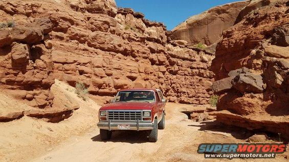 Red 1985 Ford Bronco driving on a dirt road through a canyon with red rock walls