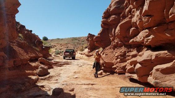 Red Ford Bronco driving on a dirt road through a canyon, spare tire mounted on the rear door