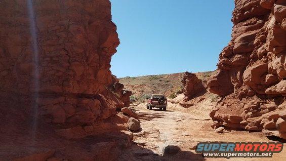 Red SUV with spare tire on rear door drives on dusty road through canyon