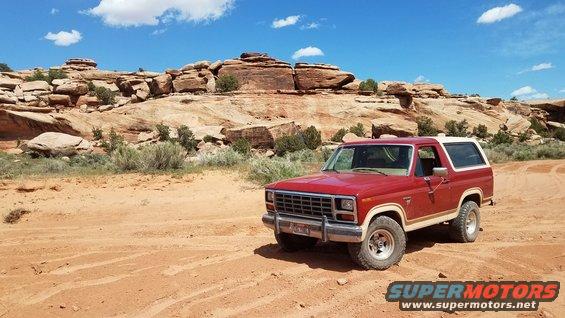 Red Ford Bronco parked on dirt road with red rock formations and blue sky in background