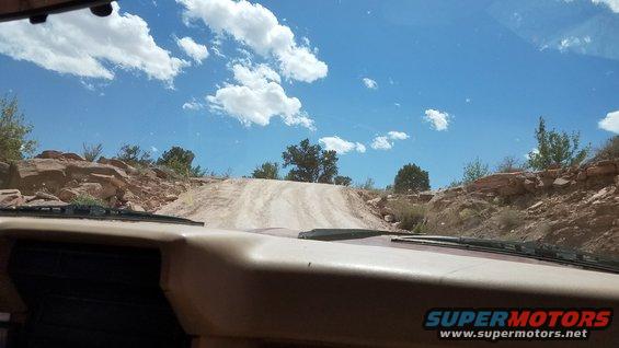 Interior view of a tan dashboard and windshield looking out at a dirt road climbing a hill in a desert