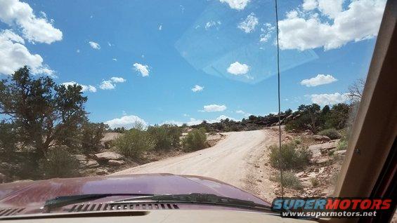 View from inside a vehicle driving on a dirt road through a desert landscape with scrub brush and rocks under a blue sky with clouds. Watermark reads SUPERMOTORS