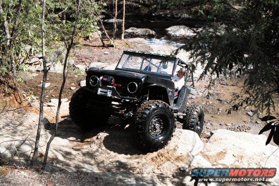 Black 1976 Ford Bronco modified for off-roading, driving over rocks near a stream