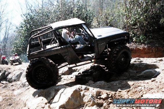 Modified off-road vehicle climbing over rocks with a person driving. Two ATVs are visible in the background