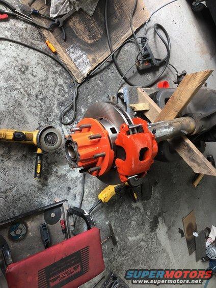 Orange brake caliper and hub assembly on a vehicle axle, with angle grinder and tools on concrete floor