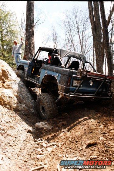 Blue 1976 Ford Bronco off-roading on a dirt trail, with a person standing on a rock nearby