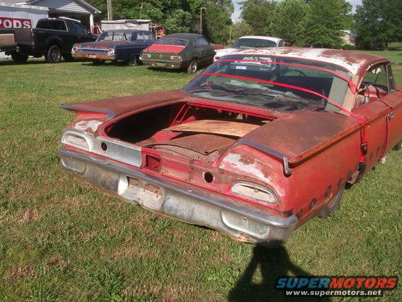 Rusty red 1960 Ford Starliner with trunk open, parked on grass. Other vehicles visible in background
