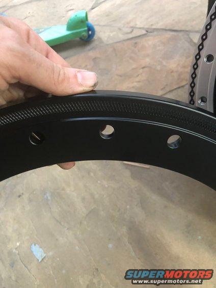Close-up of a black wheel rim with lug nut holes. A hand is holding it. A green toy scooter is in the background