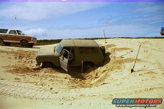 Olive green SUV stuck deep in sand dunes, driver's door open. Another SUV is parked nearby