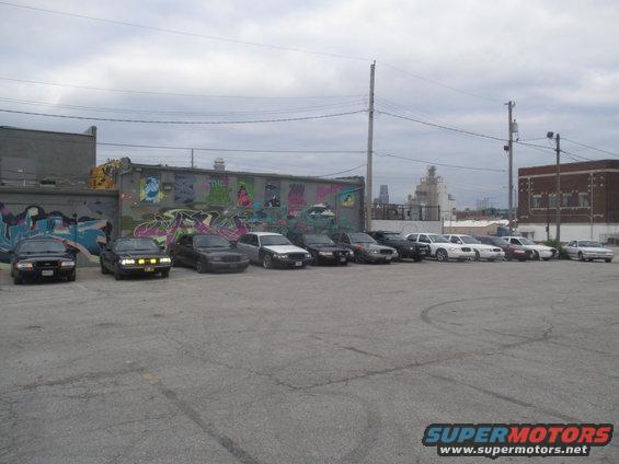 Several Ford Crown Victoria police interceptors and civilian sedans parked in a lot with graffiti-covered building in background