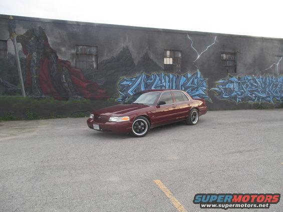 Maroon sedan parked in front of a mural, black rims, custom exhaust tips visible