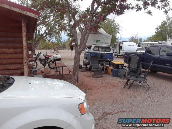 Campsite scene with a white car in foreground, motorcycle, camper van with tent, and pickup truck
