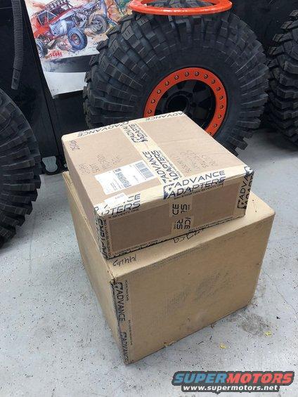 Two cardboard boxes labeled 'ADVANCE ADAPTERS' stacked on a concrete floor, with large off-road tires and a graphic of a buggy in the background