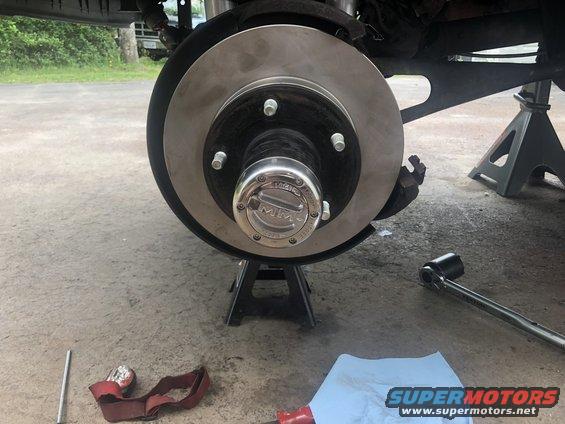 Close-up of a vehicle's front brake rotor and hub assembly, supported by jack stands. Tools are on the ground