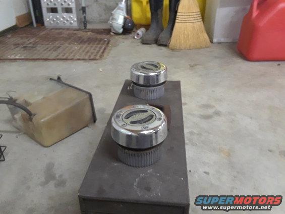 Two chrome hubcaps with black inserts sit on a dark metal plate. A plastic fluid tank is to the left