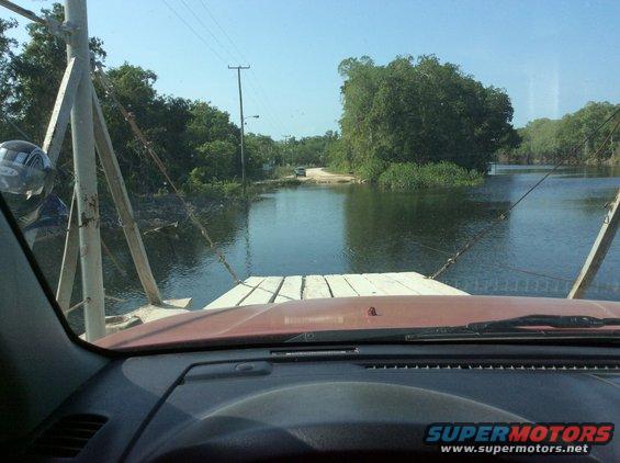 Red dashboard and windshield of a vehicle on a ferry, looking out at a water crossing with trees and a distant car
