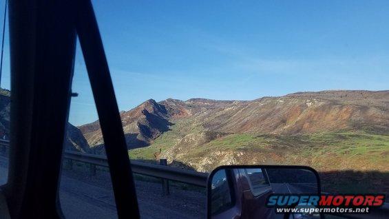 Side view mirror reflection of red truck bed and mountainous landscape