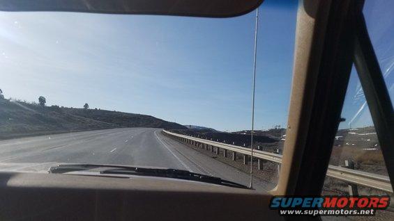 Dashboard and windshield view from inside a vehicle driving on a highway with a guardrail and rolling hills under a clear blue sky