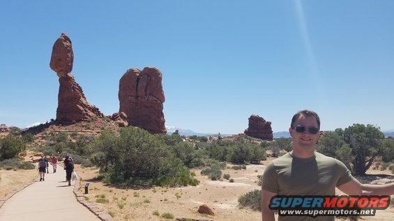 Man posing for photo with Balanced Rock and other rock formations in Arches National Park behind him