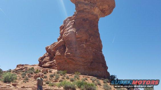 Person stands in desert landscape with large sandstone rock formation under a clear blue sky