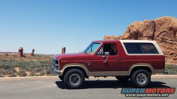 red 1985 Ford Bronco parked on road with desert rock formations in background