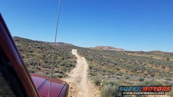 Red SUV's hood and fender visible, driving on a winding dirt road through a desert with scrub brush and distant hills under a clear blue sky