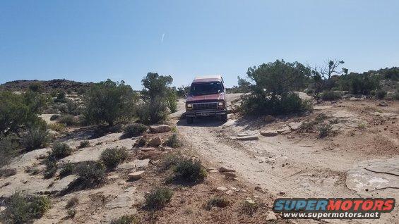 Red van with a prominent grille driving on a dirt road in a desert landscape