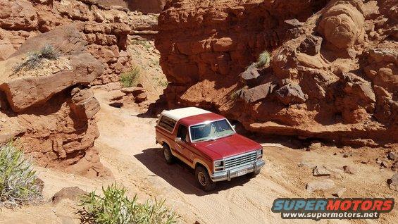 Red and white 1985 Ford Bronco driving on a dirt trail in a canyon