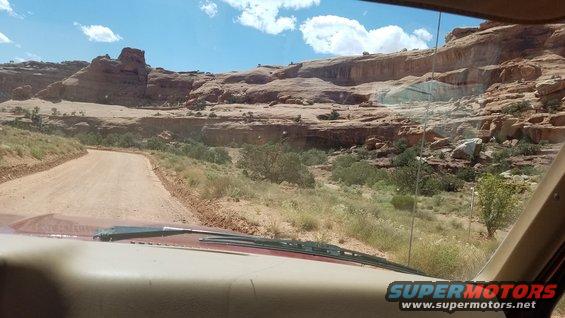 Dirt road winds through desert canyon landscape under a blue sky with clouds. View from inside a vehicle