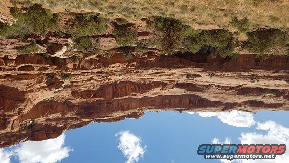 Desert canyon with red rock formations, green scrub brush, and blue sky with clouds. Watermark says SUPERMOTORS