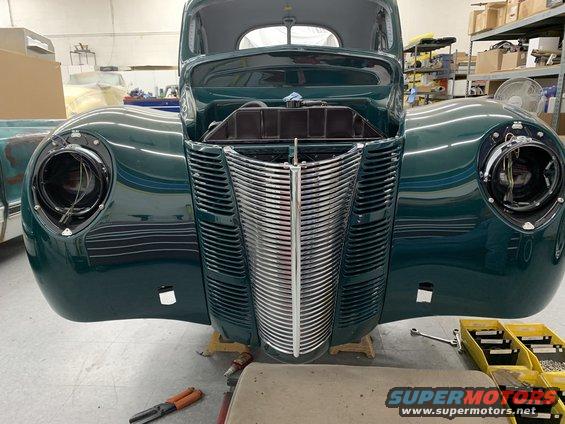 Front end of a dark green 1940 Ford Coupe with its grille and headlights visible. The car is in a workshop