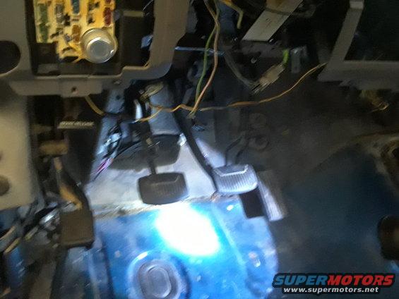 Close-up of vehicle's floorboard area showing pedals, exposed wiring, and fuse box
