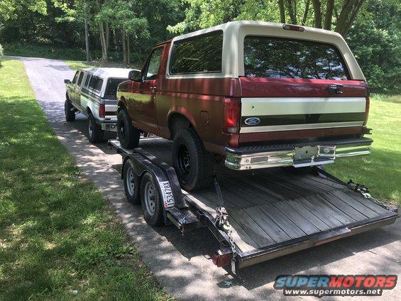 Red and tan Ford Bronco on a trailer, with another truck behind it on the road
