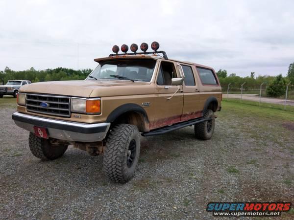 Lifted tan Ford Bronco with roof lights and side steps parked on gravel. Another vehicle is visible in the background