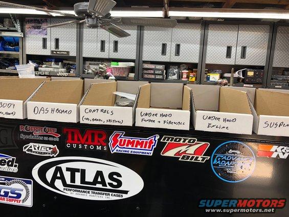 Cardboard boxes labeled 'BODY', 'DASH BOARD', 'CONTROLS', 'UNDER HOOD', and 'SUSP' on a black surface with racing stickers