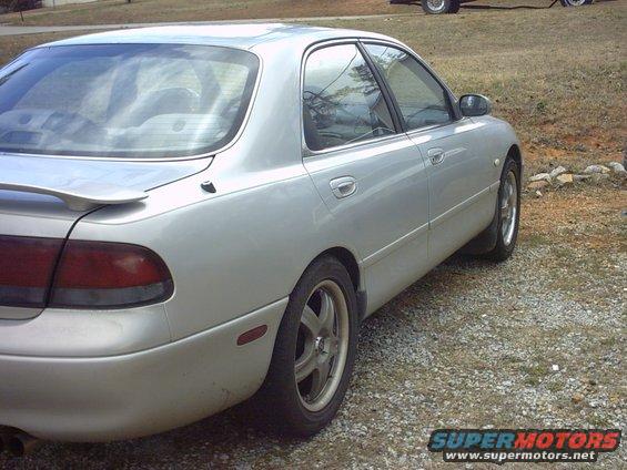 Silver sedan, rear quarter view, parked on gravel. Spoiler on trunk lid. Aftermarket wheels