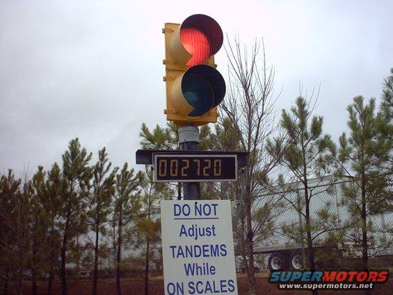 Traffic light over a digital display showing 002720, with a sign that says 'DO NOT Adjust TANDEMS While ON SCALES'. A truck trailer is visible behind trees
