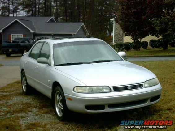 Silver 1995 Mazda 626 sedan parked on gravel, front 3/4 view. Some grass and a house in the background
