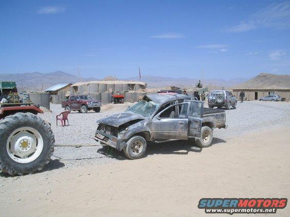Wrecked pickup truck being towed by tractor, with other vehicles and buildings in background